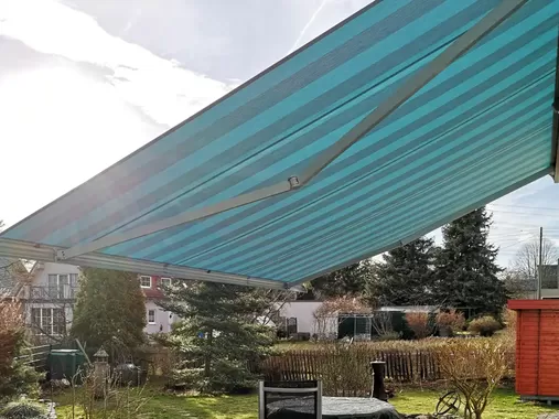 Ausgefahrene Markise mit blau gestreiftem Markisentuch spendet Schatten auf Terrasse und Gartenbereich